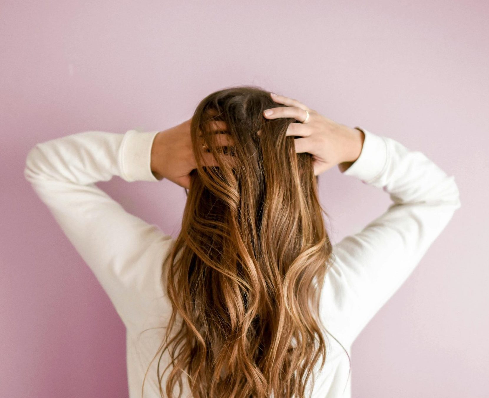 Back view of a woman with elegant long brown hair against a pink wall, showcasing stylish hair design.